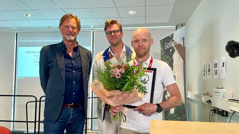Tre män står bakom en talarstol i Ljusgården på Visborg. Pristagaren Jesper håller i blommor och diplom.