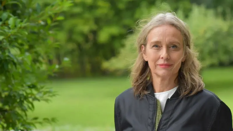 Kristina Åberg blickar leende mot kameran. I bakgrunden grönska.