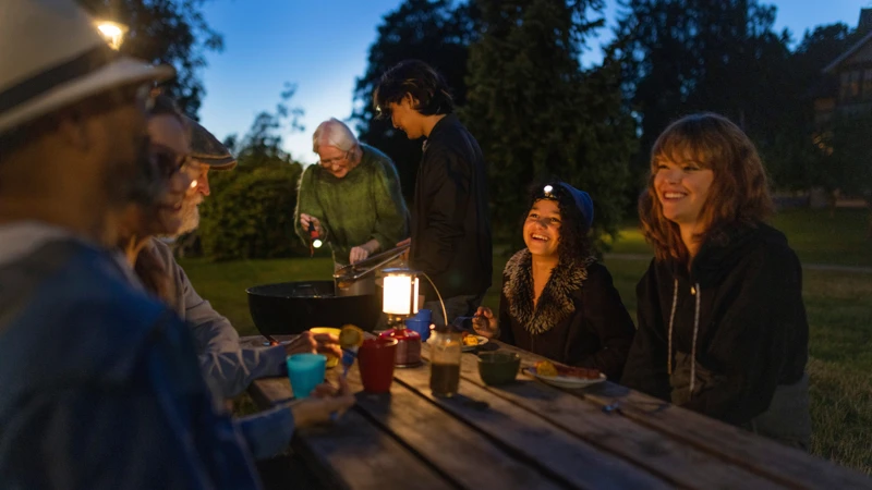 En grupp människor som sitter och skrattar runt ett träbord i en trädgård på kvällen.