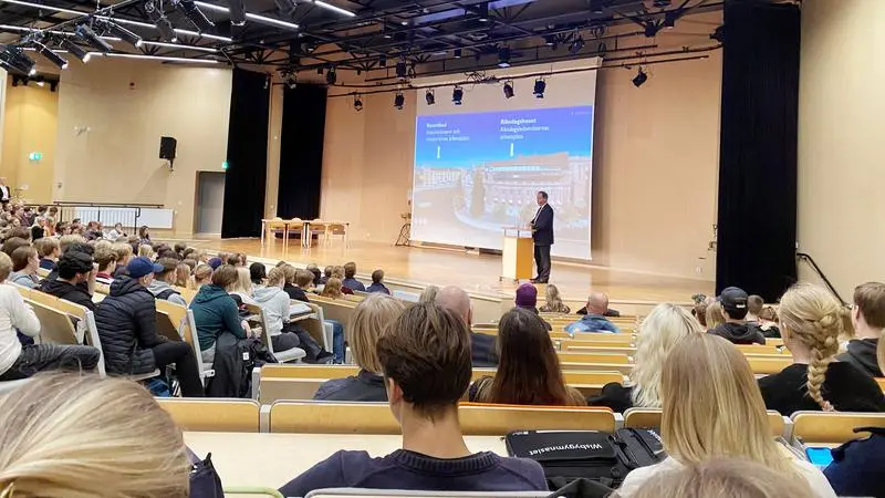 En aula full av elever som lyssnar på en talare på scen. 