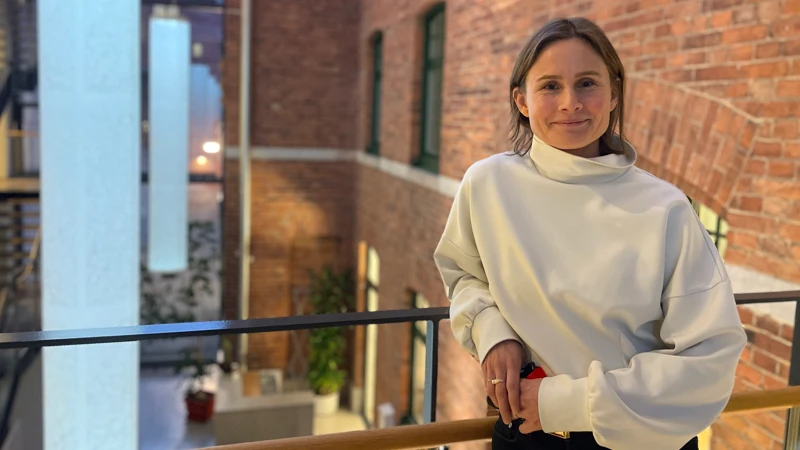 En kvinna i vit tröja står på en gångbro inomhus bredvid en tegelfasad.