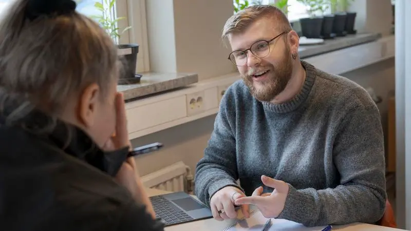 En kille i glasögon, kort hår och skägg samtalar med en kvinna som syns snett bakifrån.
