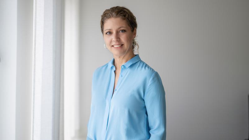 Elin Lydahl står i ett rum med blå skjorta och tittar in i kameran och ler. 