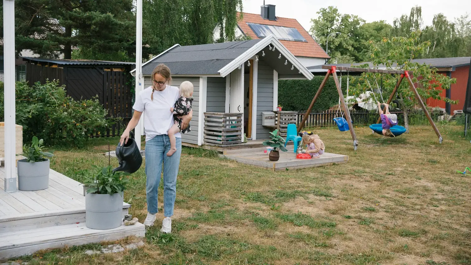 Kvinna med barn på armen vattnar i trädgården med lekande barn i bakgrunden